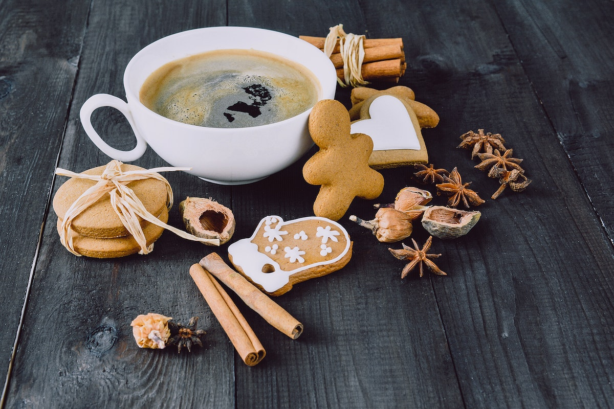 A white cup of coffee on a dark wooden table surrounded by assorted cookies, including gingerbread cookies shaped like a mitten and a person, round cookies tied with raffia, cinnamon sticks, star anise, and hazelnuts.