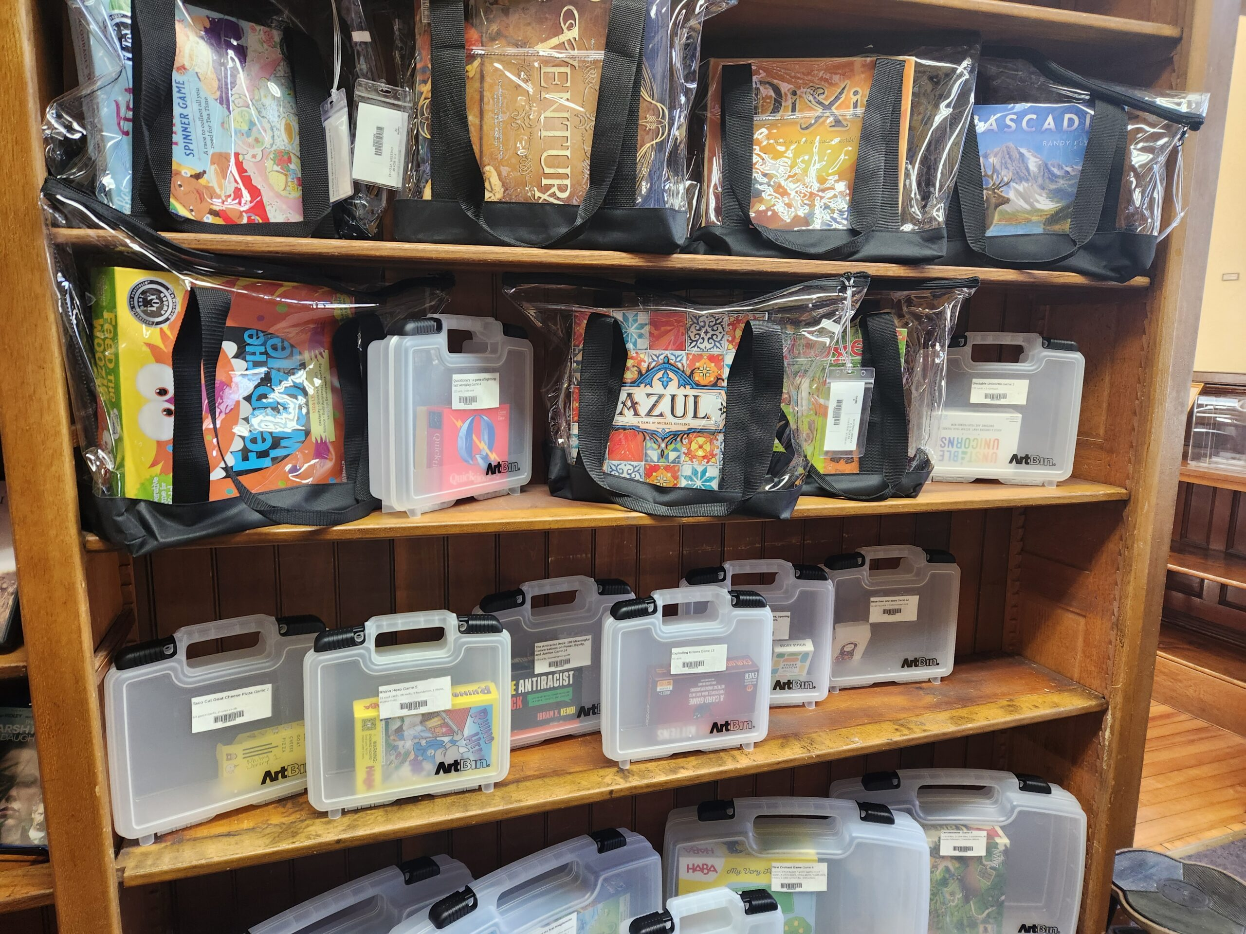 Wooden library shelves displaying various board games and kits. The upper shelves hold clear tote bags with board games including “Azul,” “Dixit,” “Cascadia,” “Feed the Woozle,” and “Zenturtle.” The lower shelves have translucent plastic carrying cases labeled “ArtBin,” each containing smaller games or kits with visible barcodes and labels. The floor is wooden, and part of the library interior is visible to the right.