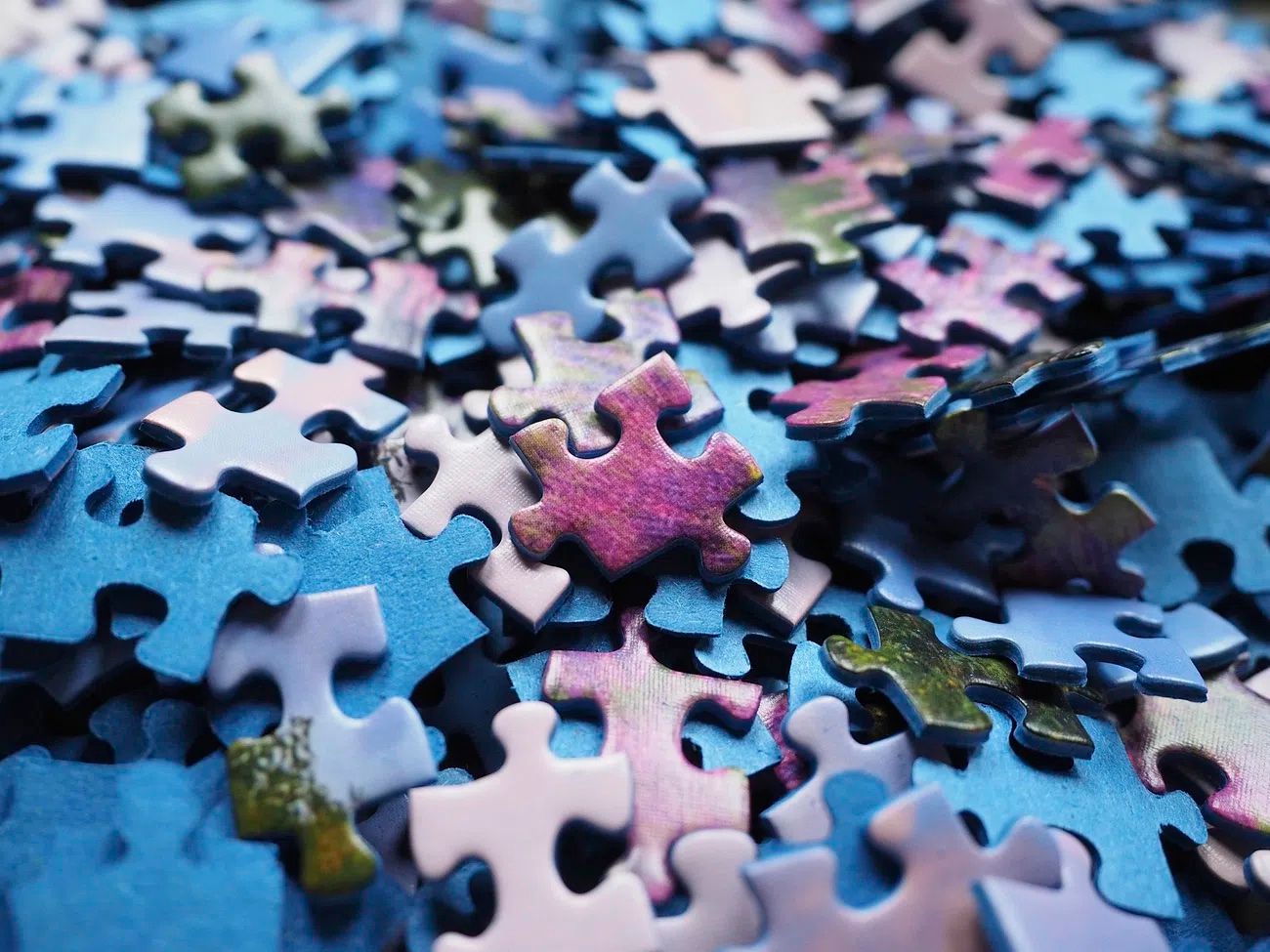 Close-up view of a pile of colorful jigsaw puzzle pieces, mostly in shades of blue, purple, and pink. The pieces are scattered randomly, showing various interlocking shapes and edges.