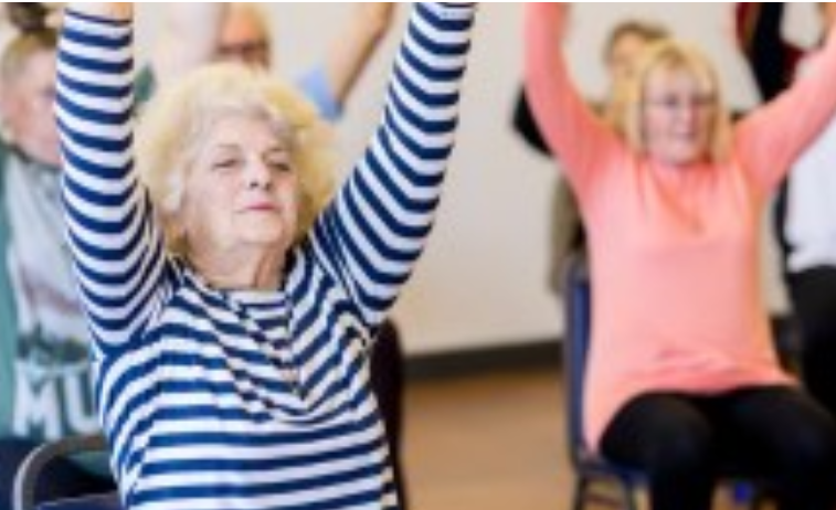 Older adults participating in a chair yoga class. A woman in the foreground wearing a blue and white striped shirt sits on a chair with her arms raised overhead. Other participants are seated behind her, also lifting their arms.