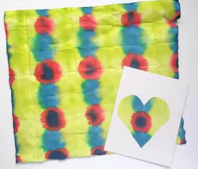 A colorful piece of Japanese tie-dyed paper featuring a repeating pattern of red, blue, and green circular designs on a yellow background. Next to it is a white card with a heart-shaped cutout, showing part of the same tie-dye pattern through the shape.