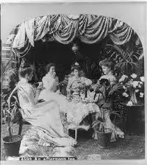 A black and white photograph showing four elegantly dressed women gathered at a small table for tea under a draped canopy, surrounded by numerous potted plants.